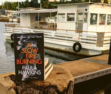 A book perched in front of a barge. The title reads 'A slow fire burning' by Paula Hawkins